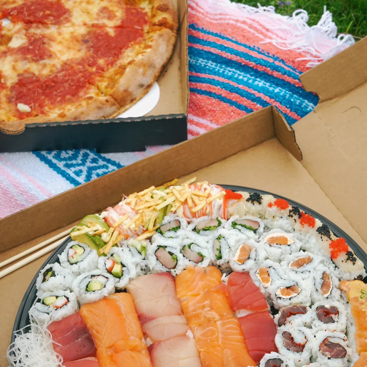 A box of assorted sushi rolls with sashimi in front of a pizza in a separate box, placed on a colorful striped cloth.