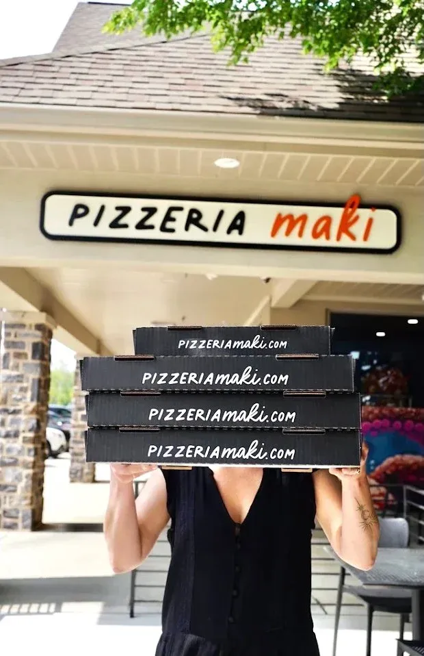 Person holding four pizza boxes in front of a pizzeria storefront with a sign that reads 'Pizzeria Maki' and the website 'pizzeriamaki.com'