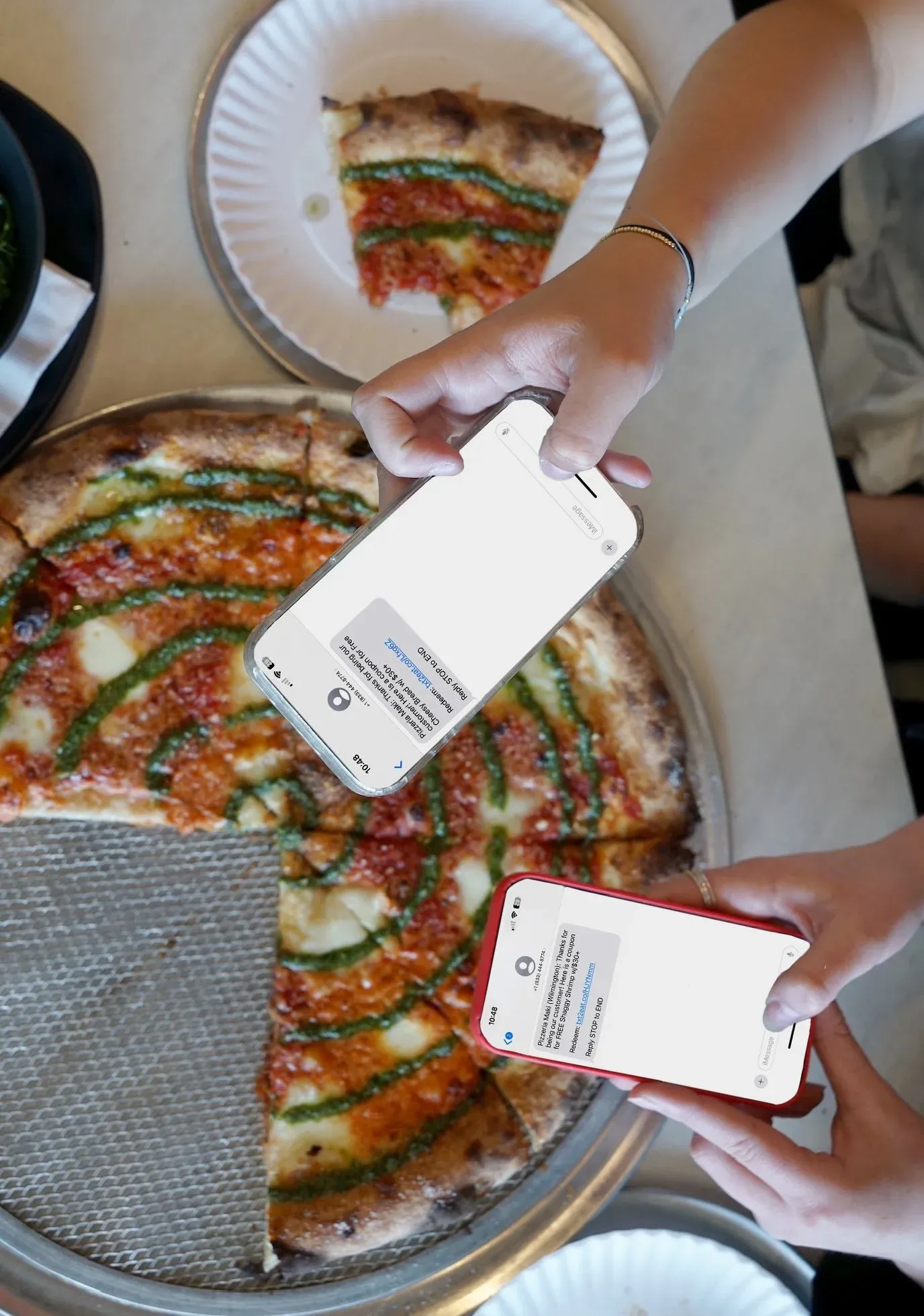 Two people holding smartphones over a partially eaten pepperoni and cheese pizza with green sauce drizzle, on a metal tray. One has a red phone, the other a white phone. A small slice of pizza on a paper plate is visible.